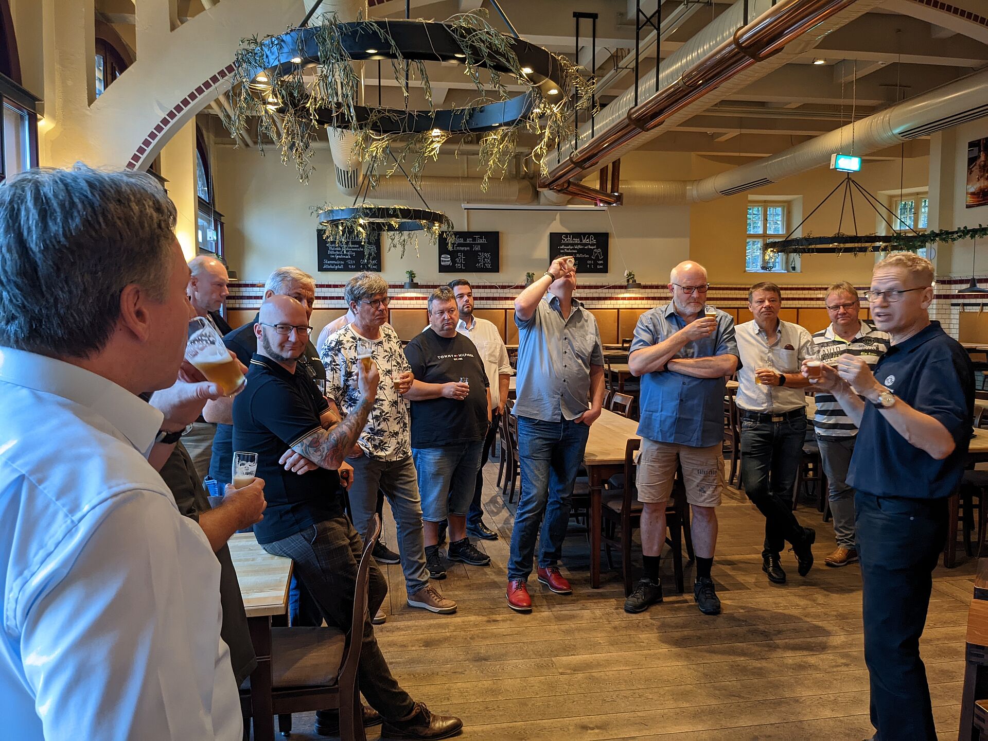 A group of people stands in a restaurant, engaged in conversation. One person is speaking to the group while others listen attentively, some holding drinks.