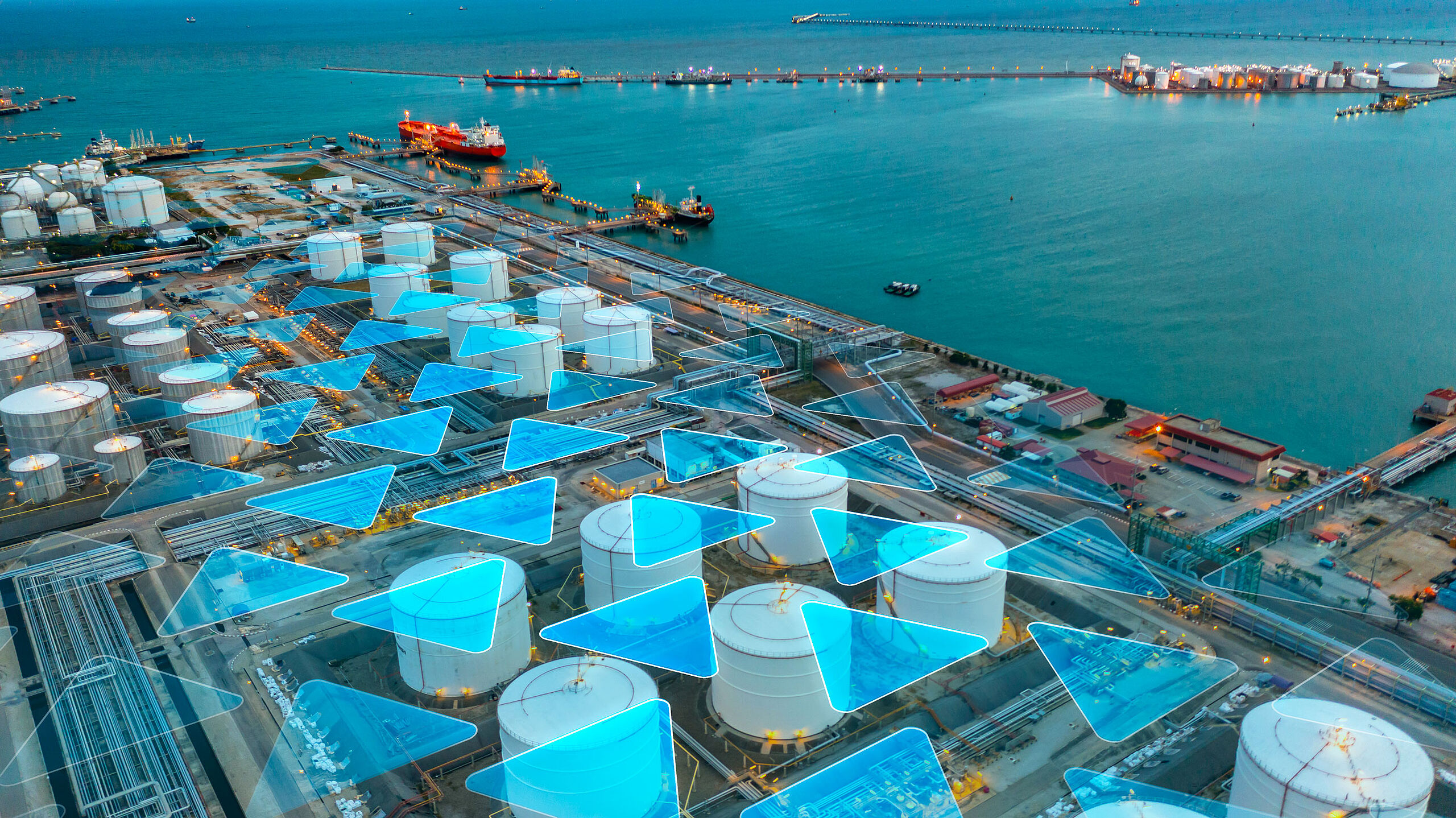 Aerial view of a coastal industrial area with large storage tanks, ships in the water, and a vibrant blue sea. Some tanks are overlaid with blue graphics.