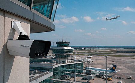 A security camera mounted on a tower overlooks an airport, with airplanes on the runway and one taking off in the background against a blue sky.