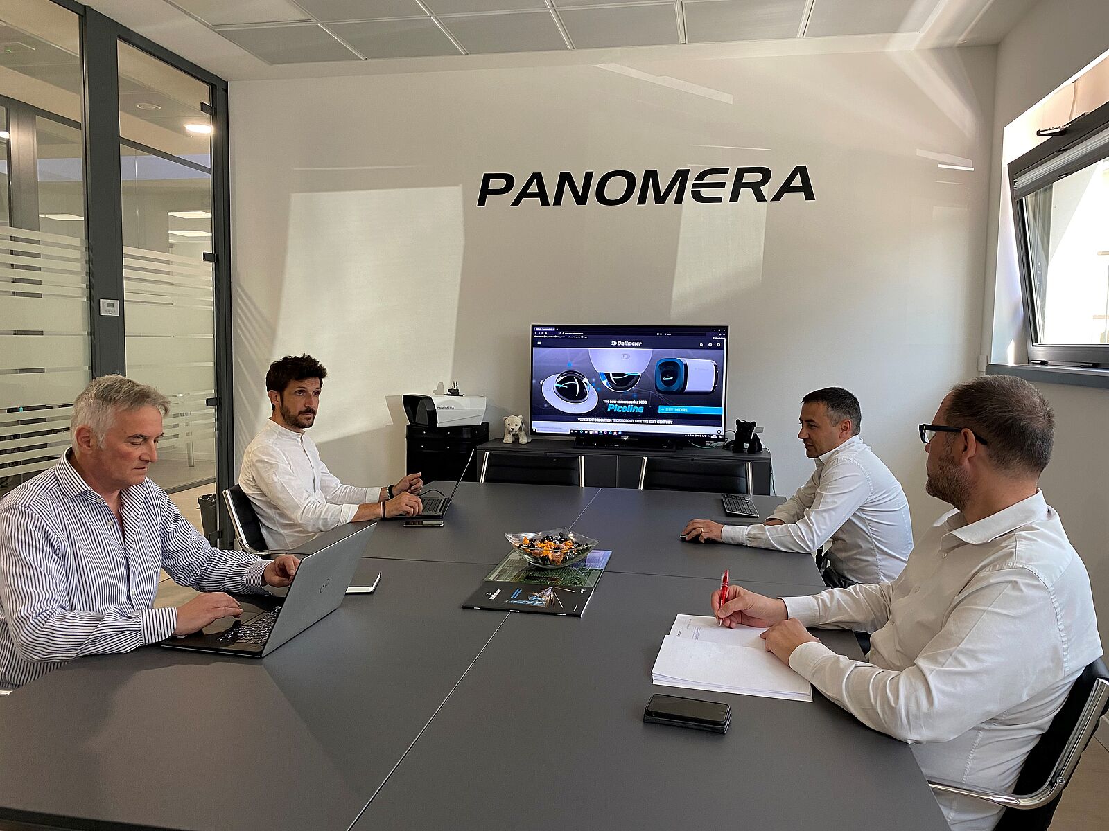 A meeting room with four men seated around a table, working on laptops. A screen displays visuals related to "PANOMERA." The atmosphere is professional and collaborative.