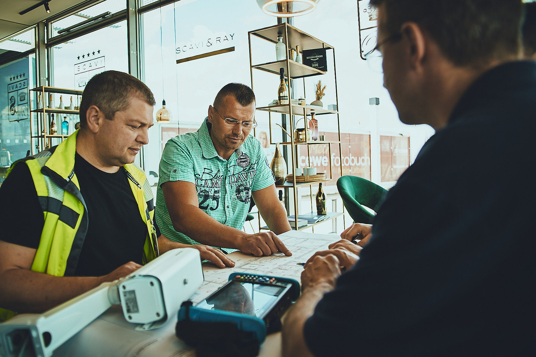 Three men are seated around a table, examining documents and plans. One is wearing a safety vest while the others are engaged in discussion, with a tablet and tools nearby.