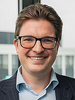 A smiling man with short, light brown hair and glasses, wearing a dark blazer over a light blue shirt, stands in front of a modern building.