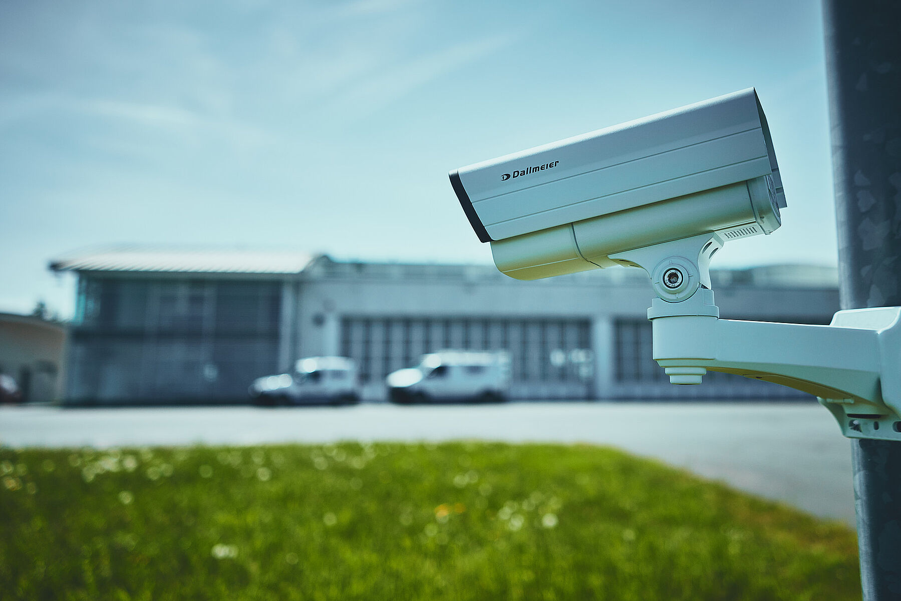 A security camera mounted on a pole, focused on a building in the background, with an open area of grass in the foreground.
