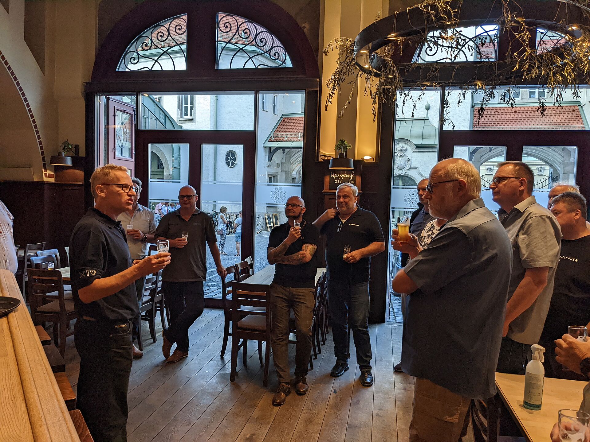 A group of men in casual attire stand in a pub, some holding drinks, as one man speaks to them. The setting includes large windows revealing an outdoor view.