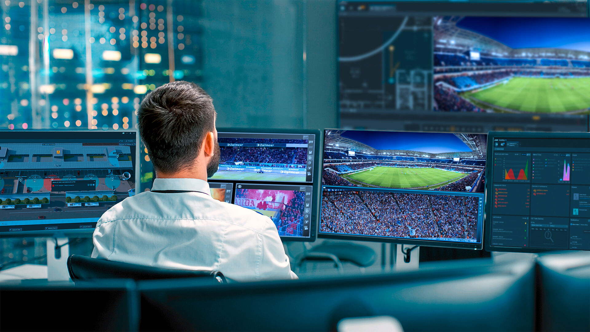 A person wearing a white shirt is seated in front of multiple computer monitors displaying sports data and live footage of a stadium, with city lights in the background.