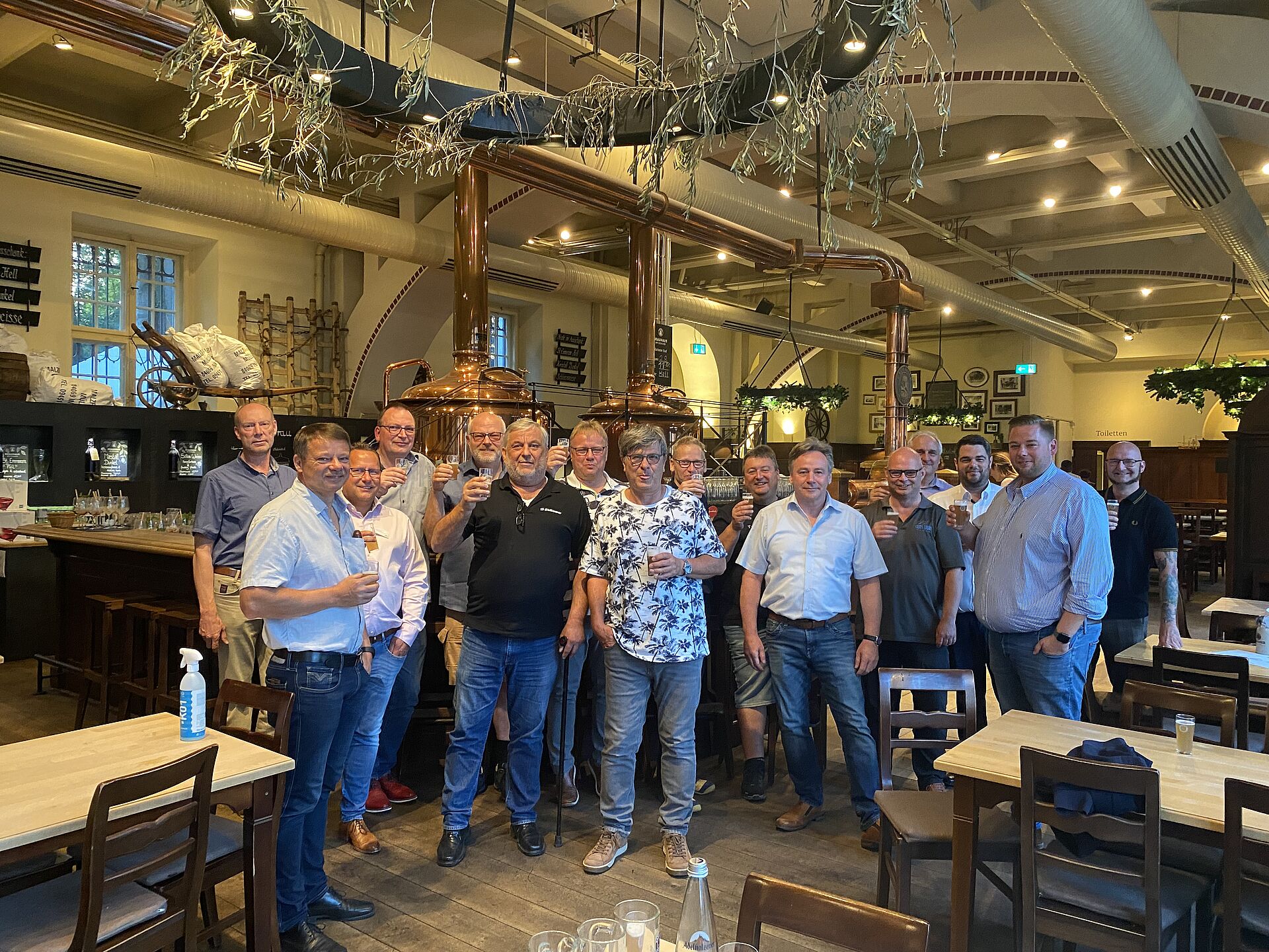 A group of people stands together in a brewery, smiling and holding drinks, with brewing equipment visible in the background. Tables and chairs are arranged around them.