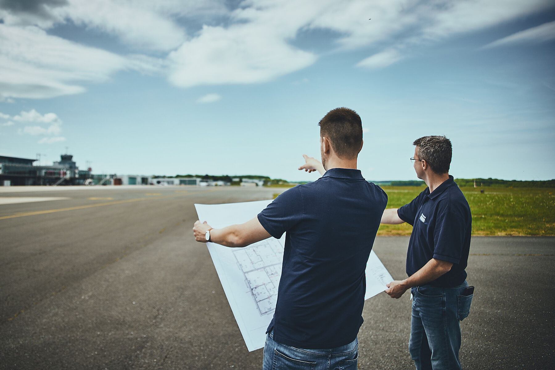 Two men, standing on an airfield, look at a blueprint and point towards the runway with an airport in the background under a partly cloudy sky.
