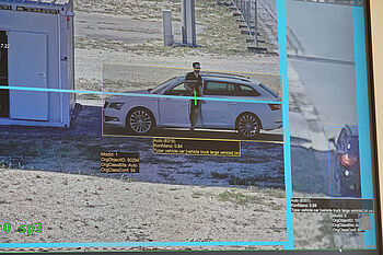 A surveillance screen captures a white vehicle parked next to a trailer, with a person standing beside it, highlighted with tracking information.