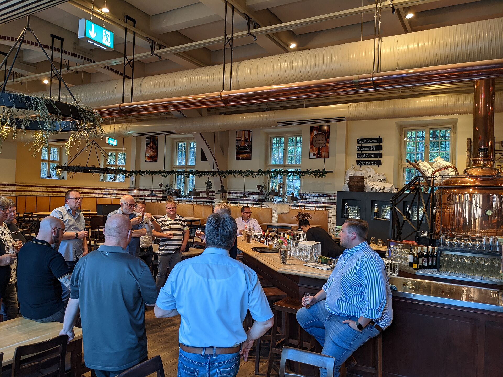 A group of people stands around the bar in a bustling establishment, engaged in conversation, with brewing equipment visible in the background.