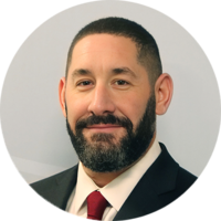 Headshot of a man with a beard, wearing a black suit and a red tie, smiling against a neutral background.