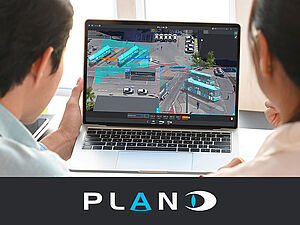 Two people are sitting closely together, viewing a laptop screen displaying a monitoring interface with images of a transportation area, featuring buses and vehicles. The logo "PLAN" is visible at the bottom.