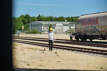 A railway worker in a yellow hard hat stands on train tracks, holding a clipboard, with a train and industrial buildings in the background.