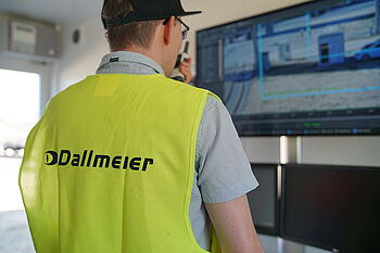 A person wearing a yellow vest with the text "Dallmeier" on the back is standing in front of a large monitor displaying video footage.