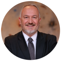 A smiling man with a beard, wearing a black suit and tie, poses confidently against a blurred background.