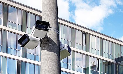 Three security cameras are mounted on a concrete pole, overlooking a modern building with large glass windows and a blue sky in the background.