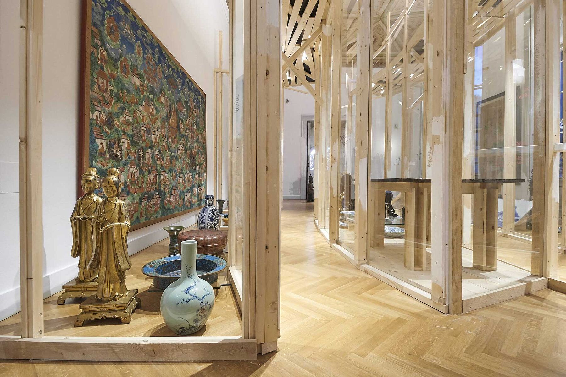 Interior view of an art exhibition featuring wooden structures, a large tapestry on the wall, and several decorative vases and statues displayed on the floor.