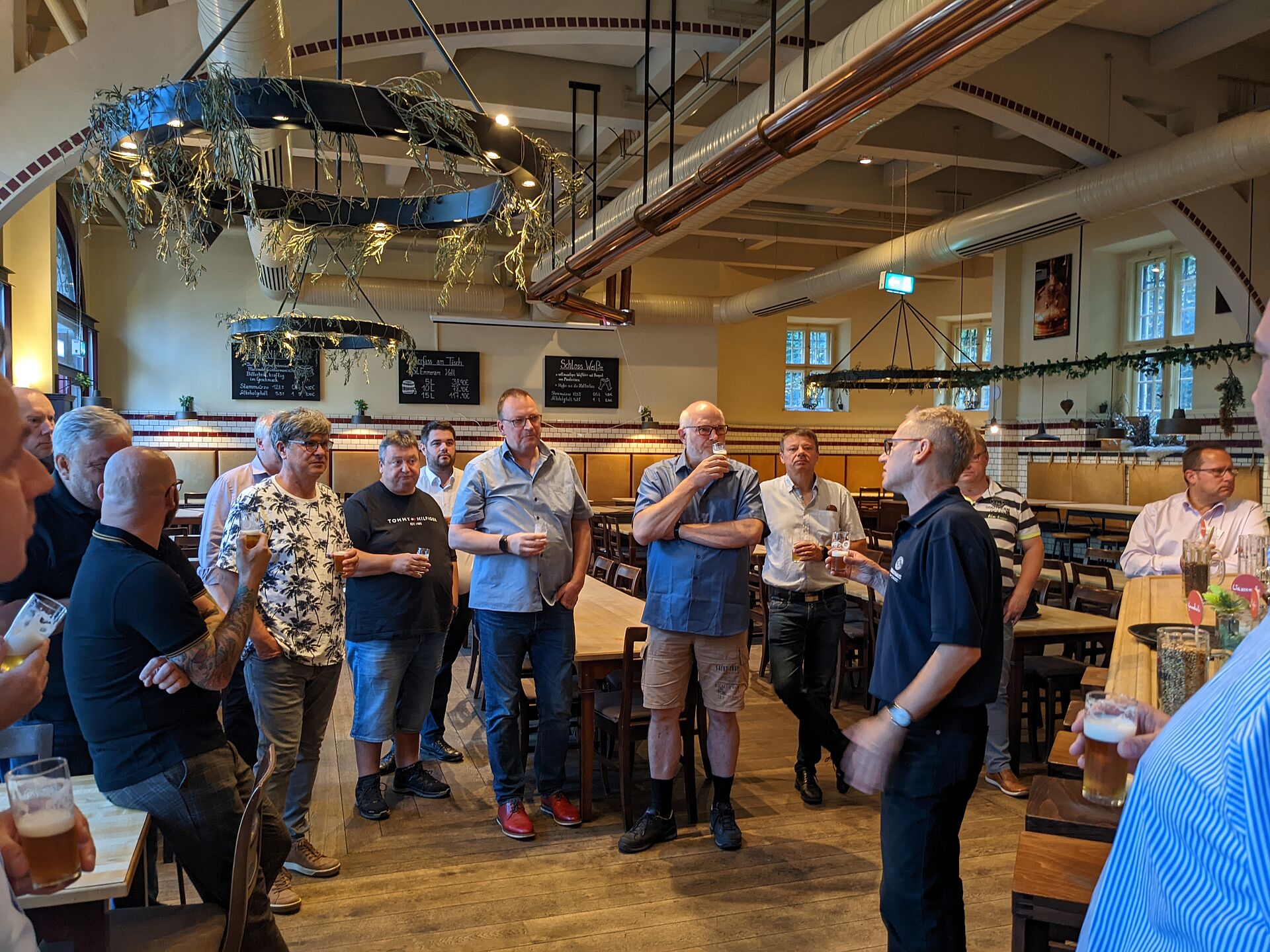 A group of men stands in a casually decorated restaurant, listening to a speaker while holding drinks, engaged in conversation.