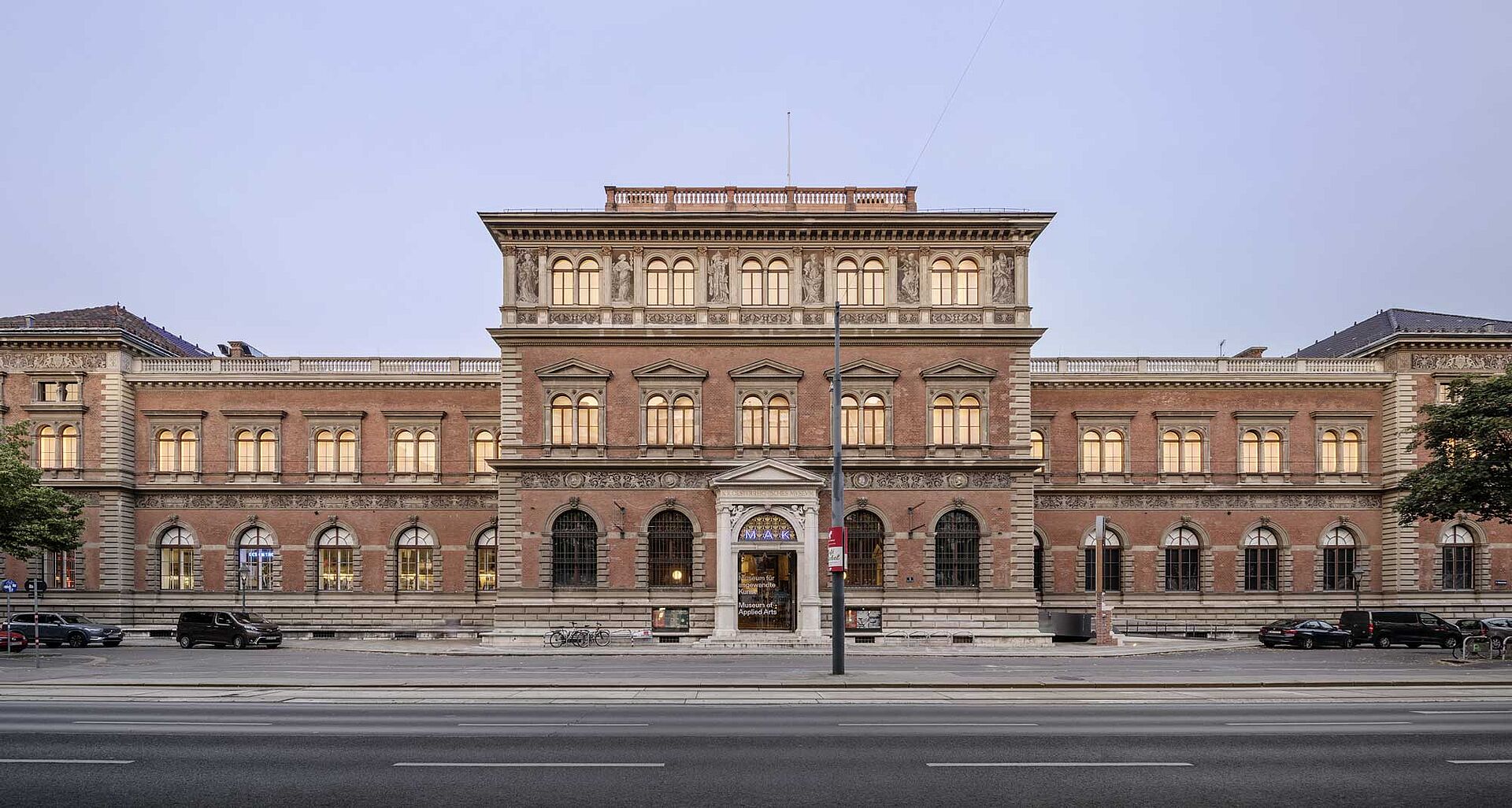 Historic building with ornate architecture, featuring large windows and decorative elements, viewed from the street. Trees and parked bicycles are visible in the foreground.