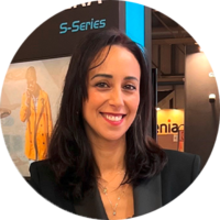 A woman with shoulder-length dark hair smiles in front of a display featuring the text "S-Series." She wears a black blazer and has a necklace.