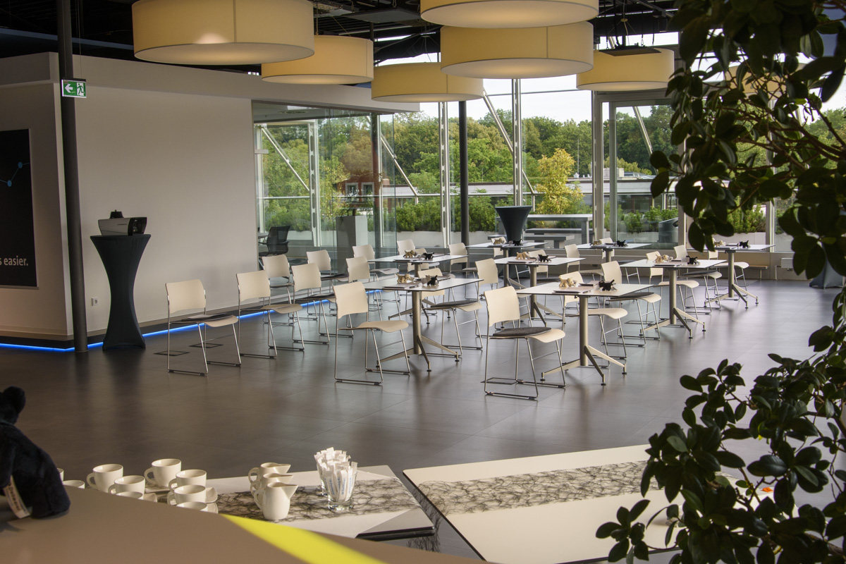 Modern dining area with white chairs and tables, large windows showing greenery outside, and pendant lights hanging from the ceiling. A small serving table with cups is visible.