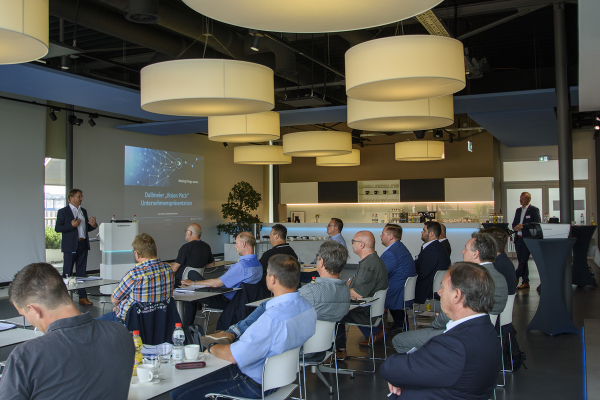 A group of people seated in a modern conference room attentively listens to a speaker presenting in front of a screen.