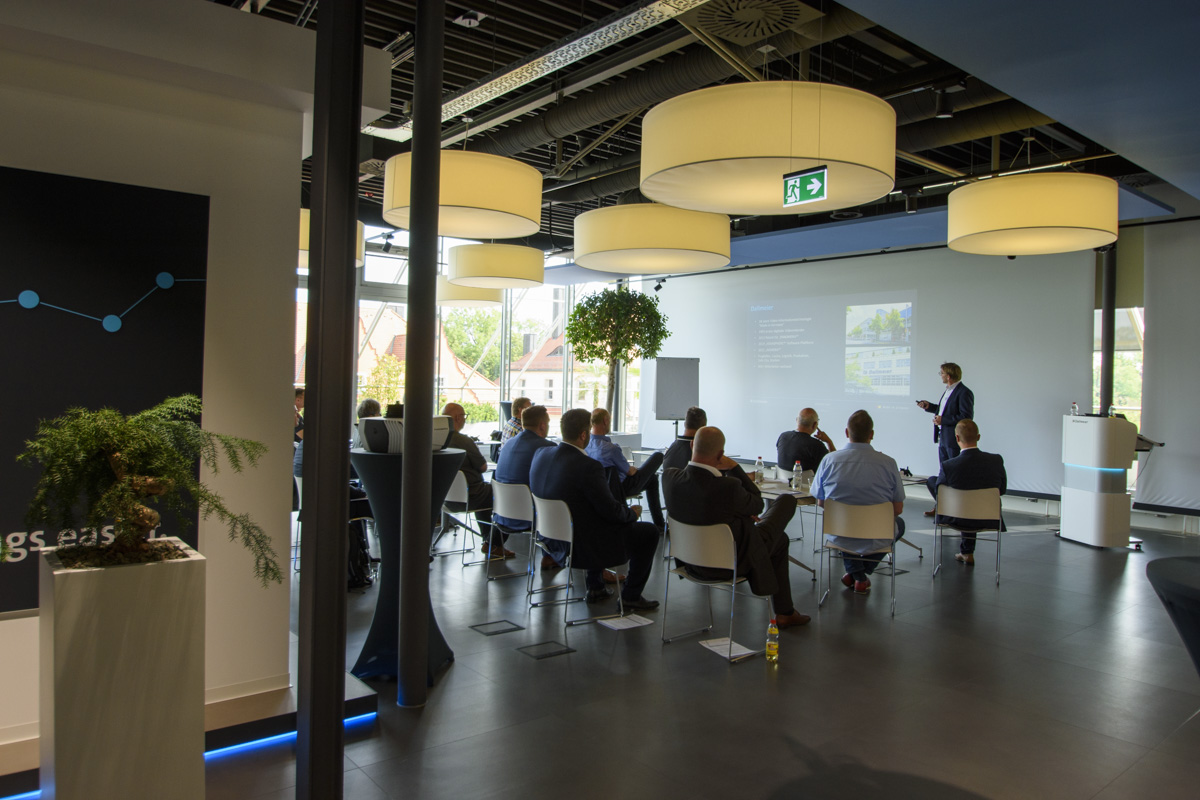 A conference room with a speaker addressing an audience seated in chairs. The room is well-lit, featuring modern decor and large windows. A presentation is displayed on a screen.