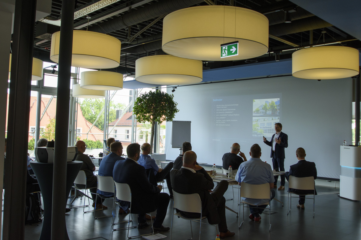 A group of people seated in chairs facing a presenter who is speaking in a bright room with large windows and modern lighting fixtures. A presentation is displayed on a screen.