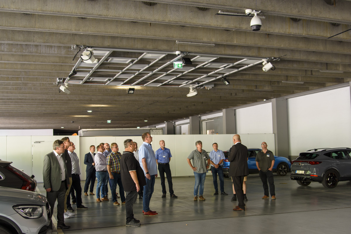 A group of people stands in a spacious, well-lit garage, attentively listening to a speaker. Several cars are visible in the background.