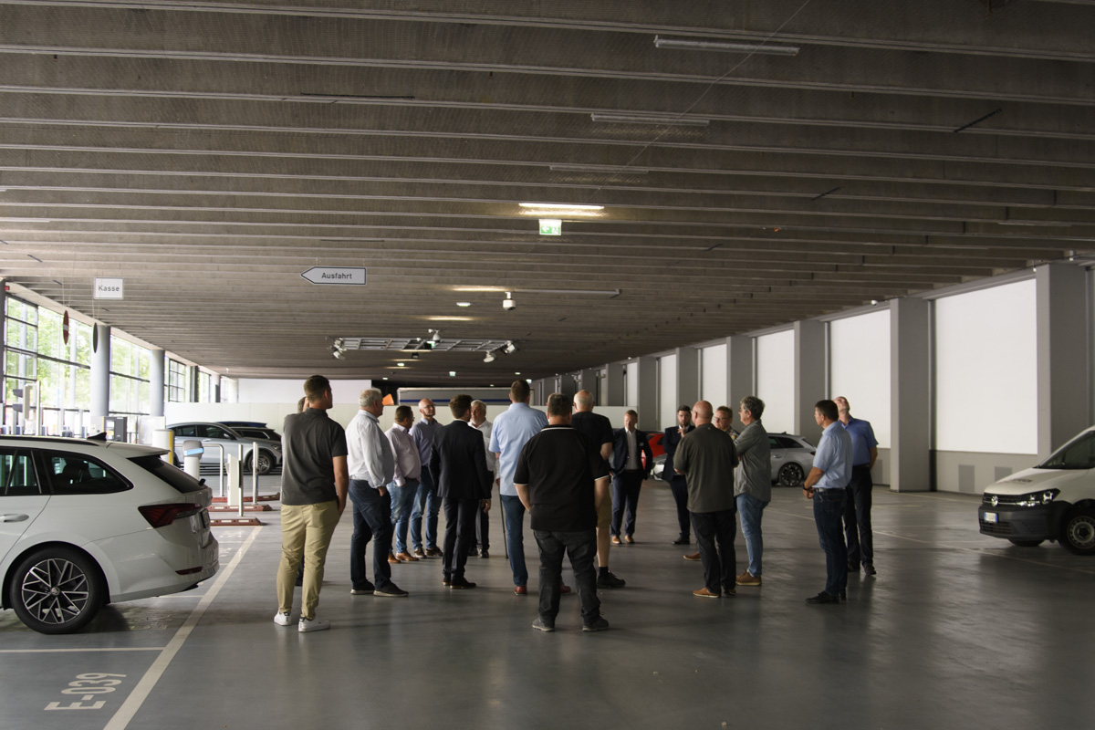 A group of people gathered in a spacious parking garage, discussing among themselves with vehicles parked in the background. The garage features concrete walls and overhead lighting.