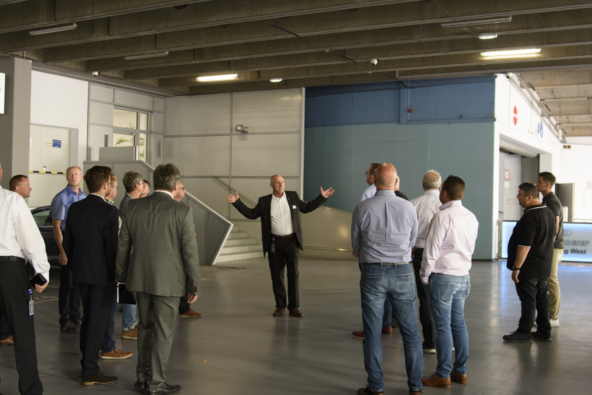 A group of professionals stands in a spacious, industrial setting, attentively listening to a speaker gesturing with hands, suggesting a discussion or presentation.