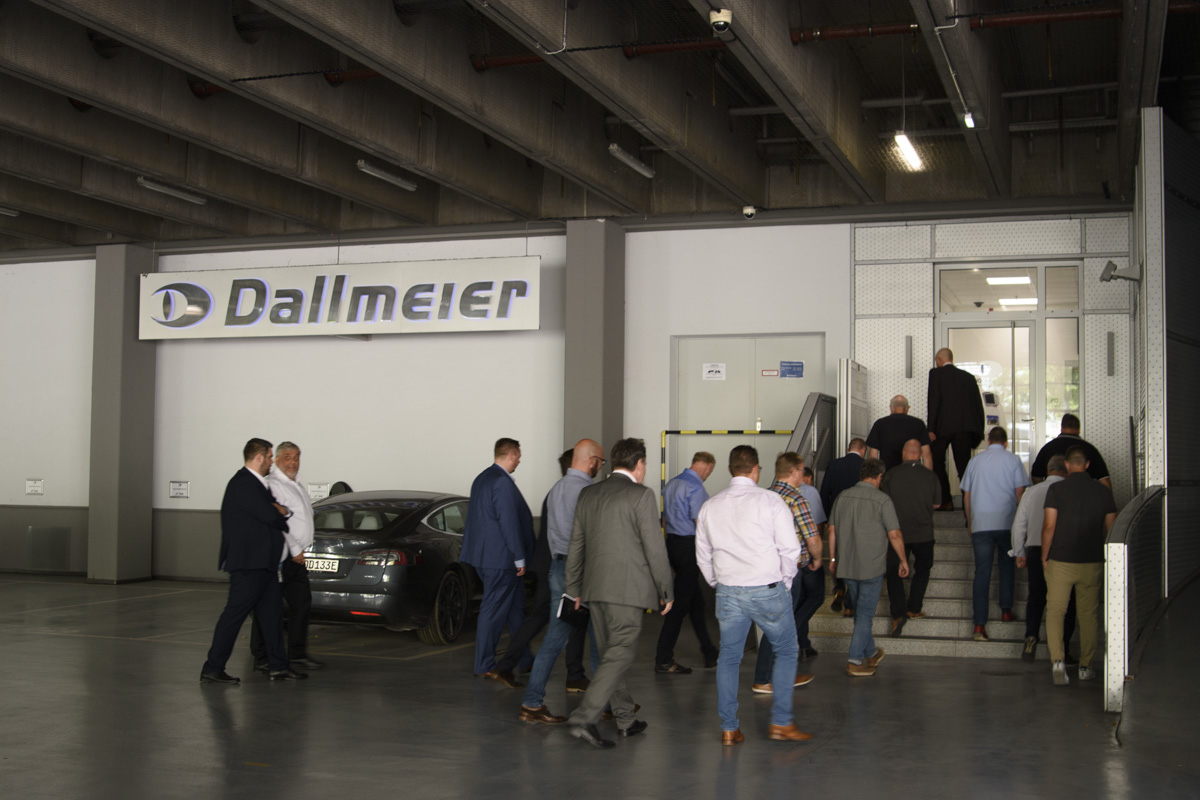 A group of people in business attire walking towards the entrance of a building with a "Dallmeier" sign above. A car is parked nearby.
