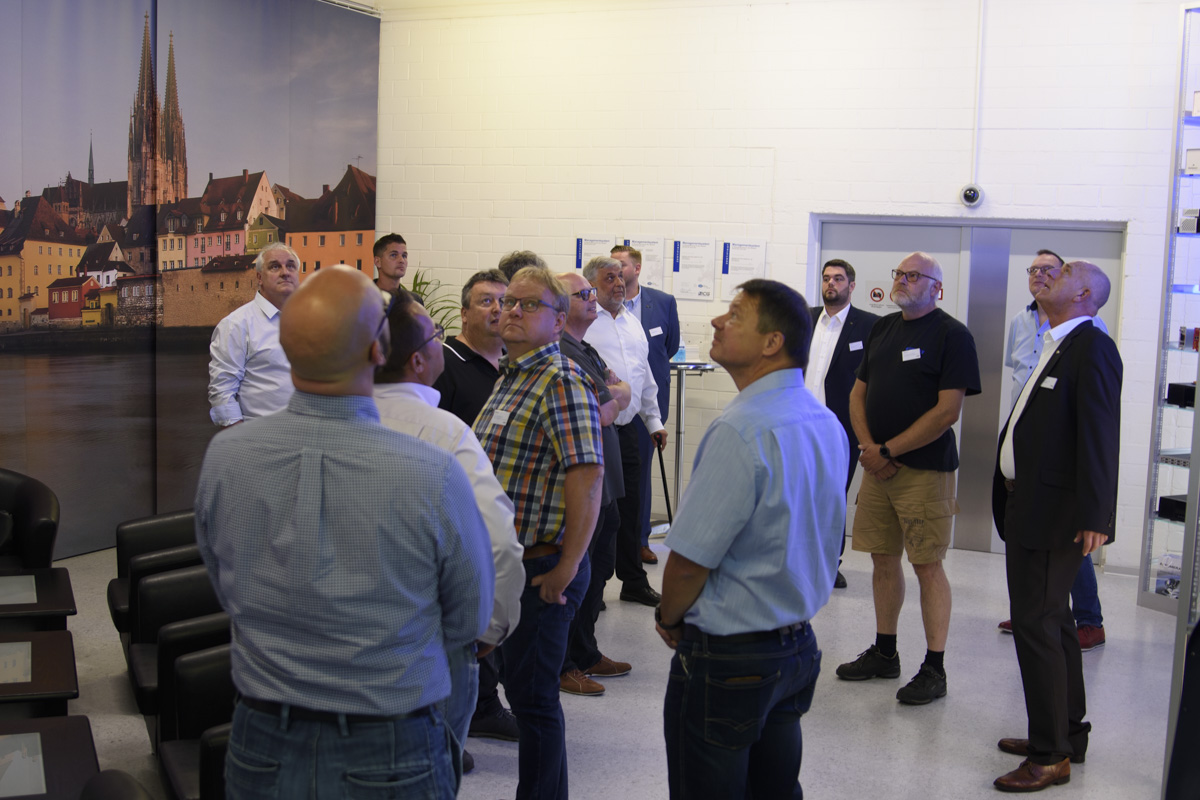 A group of men, dressed in business and casual attire, gather inside a room, looking upwards, with a large scenic mural of a city in the background.