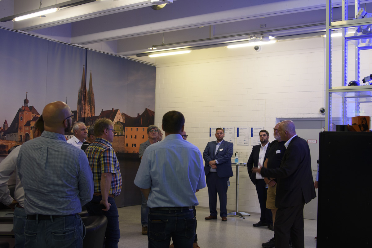 A group of people stands in a seminar setting, listening to a speaker in front of a backdrop of a cityscape. Some attendees take notes or engage in discussion.