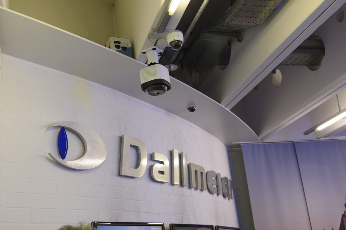 Sign for "Dallmeier" displayed on a white wall, with a surveillance camera mounted above it. The background shows gray curtains and industrial lighting.