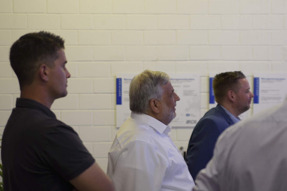 Three men are standing in profile, looking at something out of view, with a light-colored wall and documents partially visible in the background.