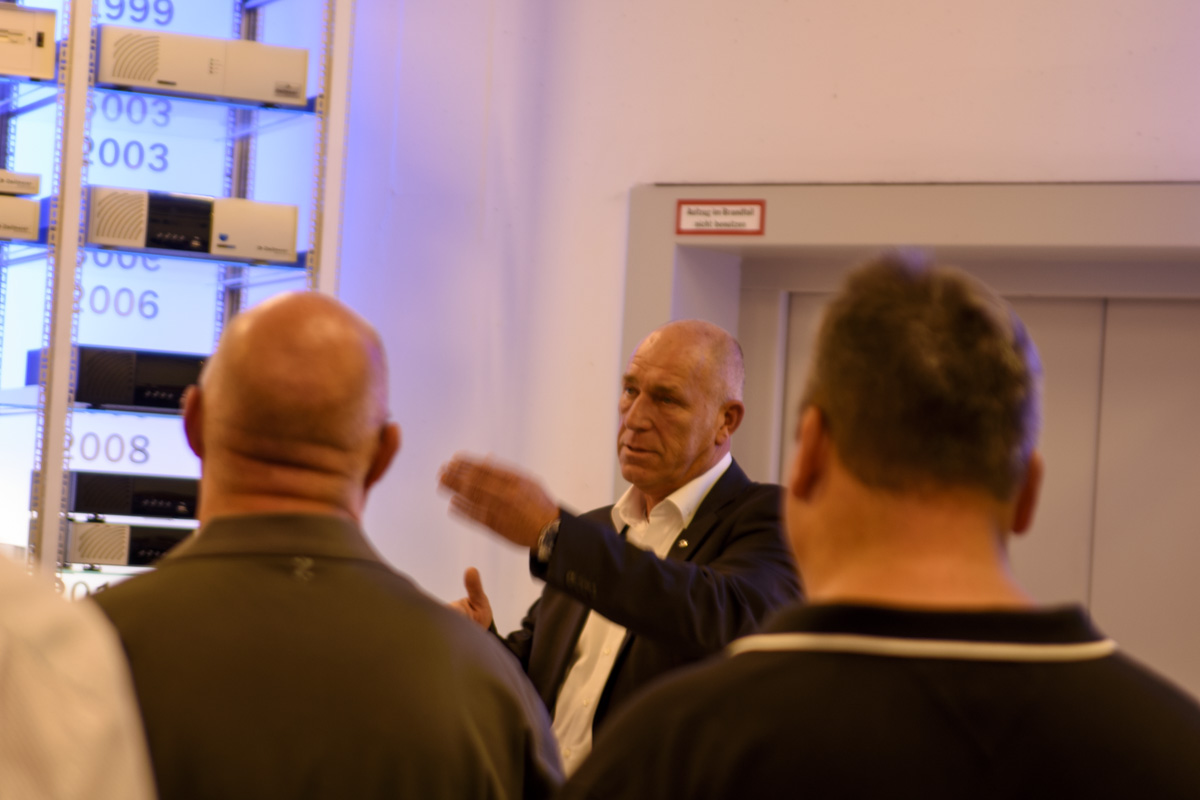 A speaker gestures while addressing a small group of people, surrounded by display panels labeled with years. The setting appears to be an informational presentation.