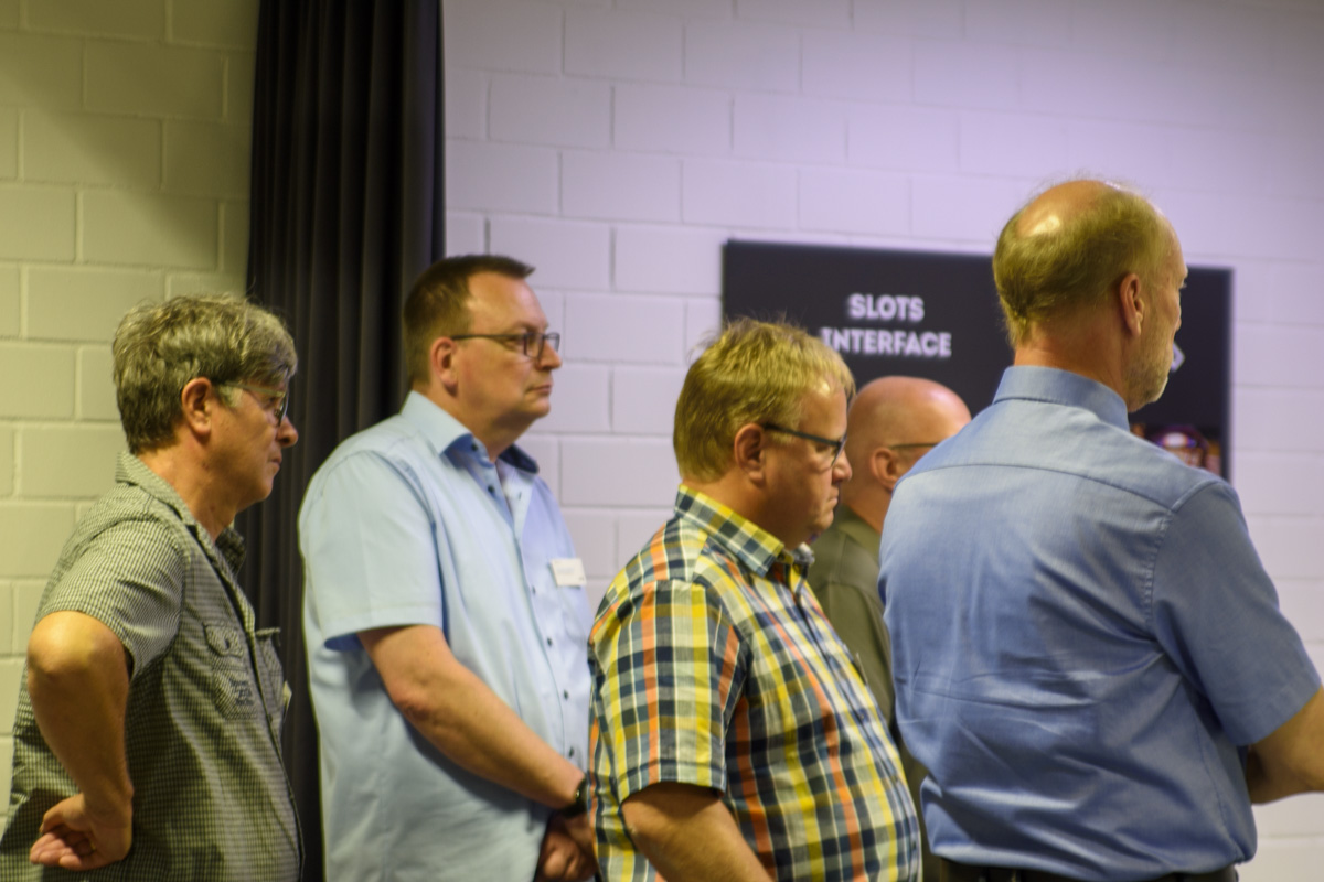 A group of five men stands in a row, facing forward, in a conference setting. They appear engaged and attentive, with various styles of shirts.