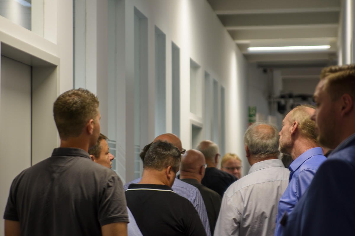 A group of men standing in a hallway, facing away from the camera, with a line forming towards an elevator.
