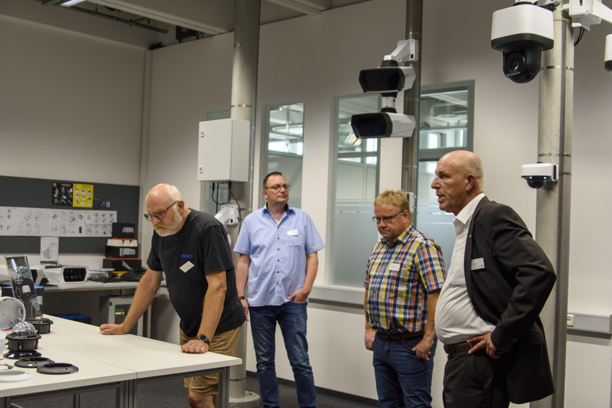 Four men are gathered around a table in an industrial setting, examining various devices and equipment, while cameras and technology are mounted nearby.