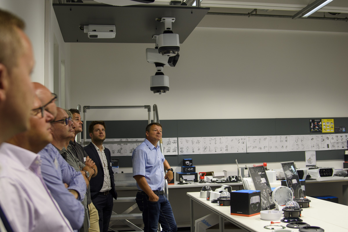 A group of people observing technology products in a modern lab setting, with displays of various devices on tables and a projector mounted on the ceiling.