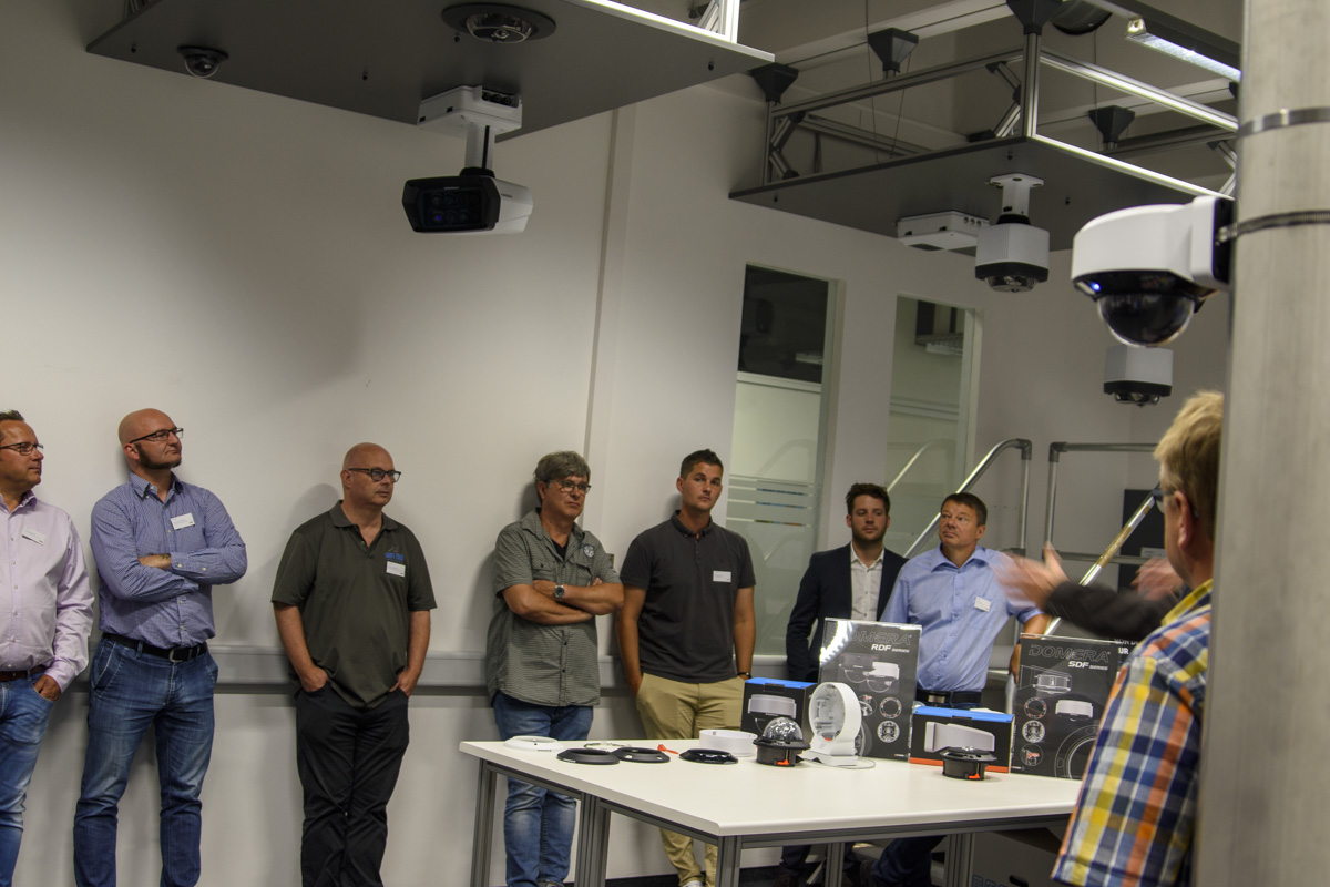 A group of men stands in a modern meeting room, listening to a speaker near a table displaying various technology products and equipment, including ceiling-mounted projectors.