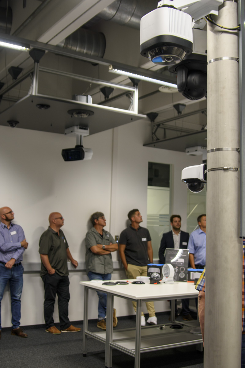 A group of people stands in a tech presentation space, observing various equipment displayed on a table, with ceiling-mounted cameras visible above.