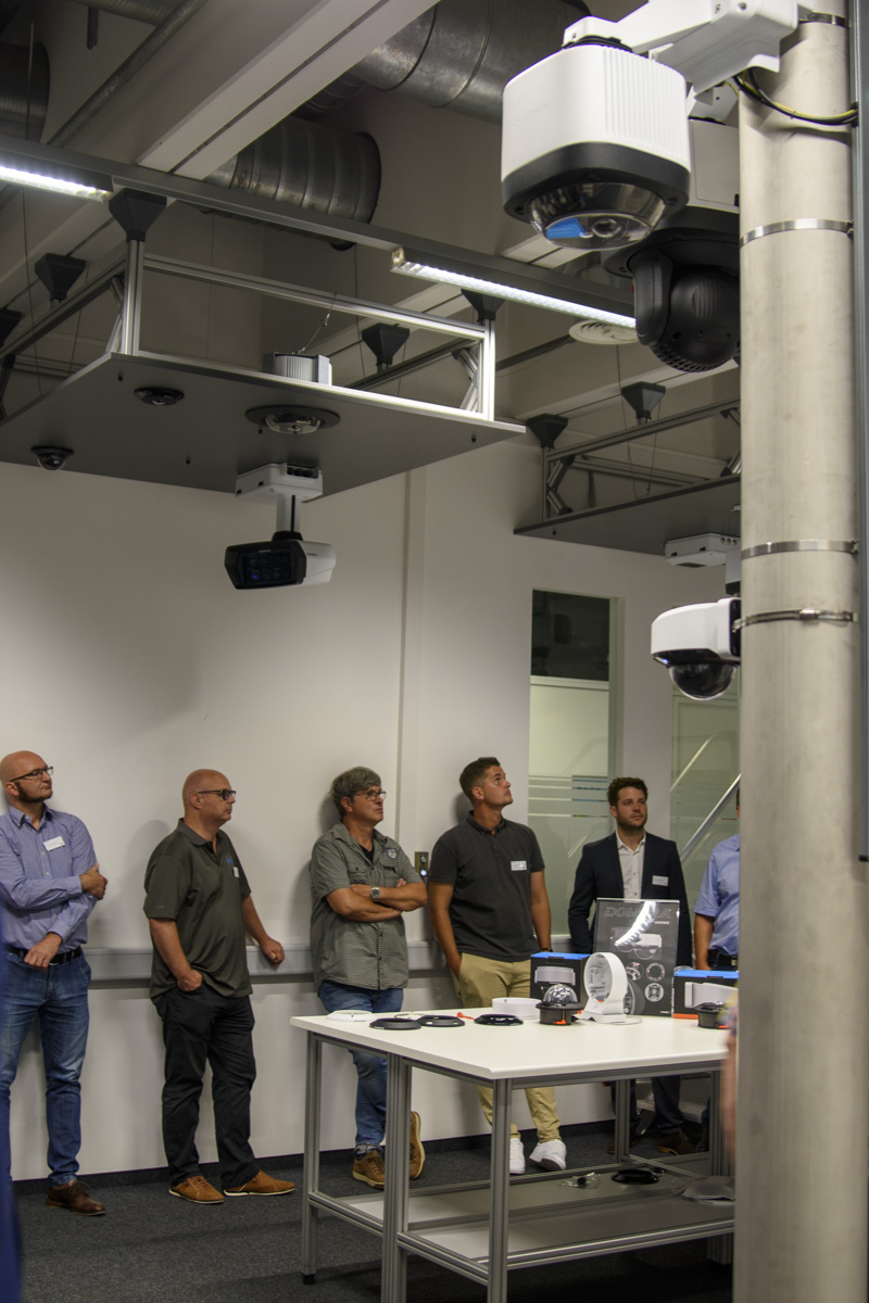 A group of six individuals stands in front of a presentation table in a modern room, with ceiling-mounted projectors and cameras visible above.