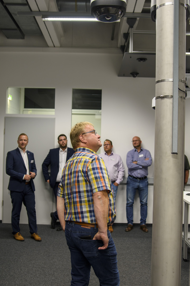 A group of men stands in a modern room, some looking toward a camera mounted on a pole, while one man in a plaid shirt gazes upward.
