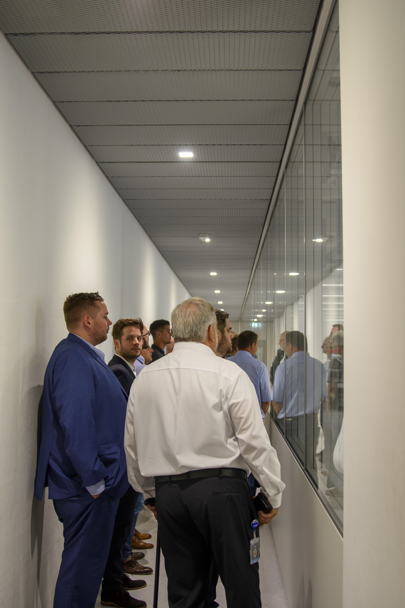 A group of men dressed in business attire stands in a narrow hallway, observing a display case on one side, illuminated by overhead lights.