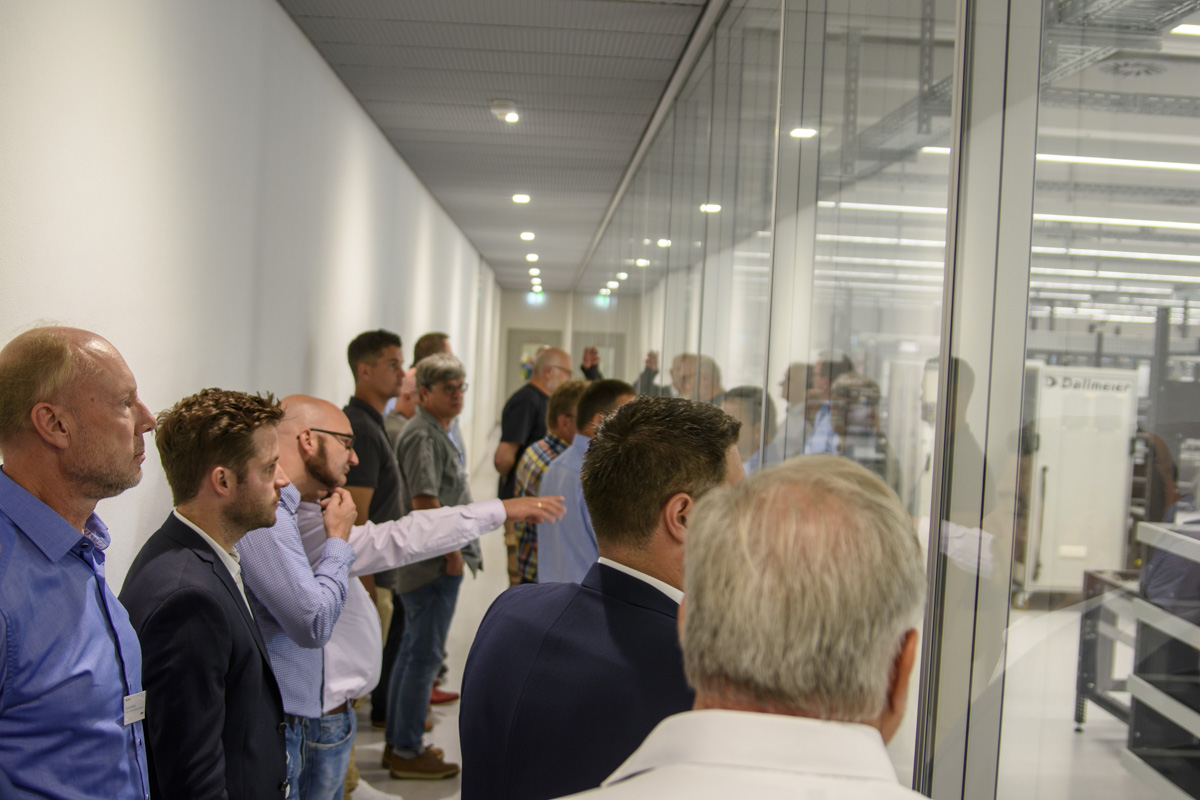 A group of people stands in a line, observing through a glass wall, likely observing machinery or a demonstration in a modern facility.
