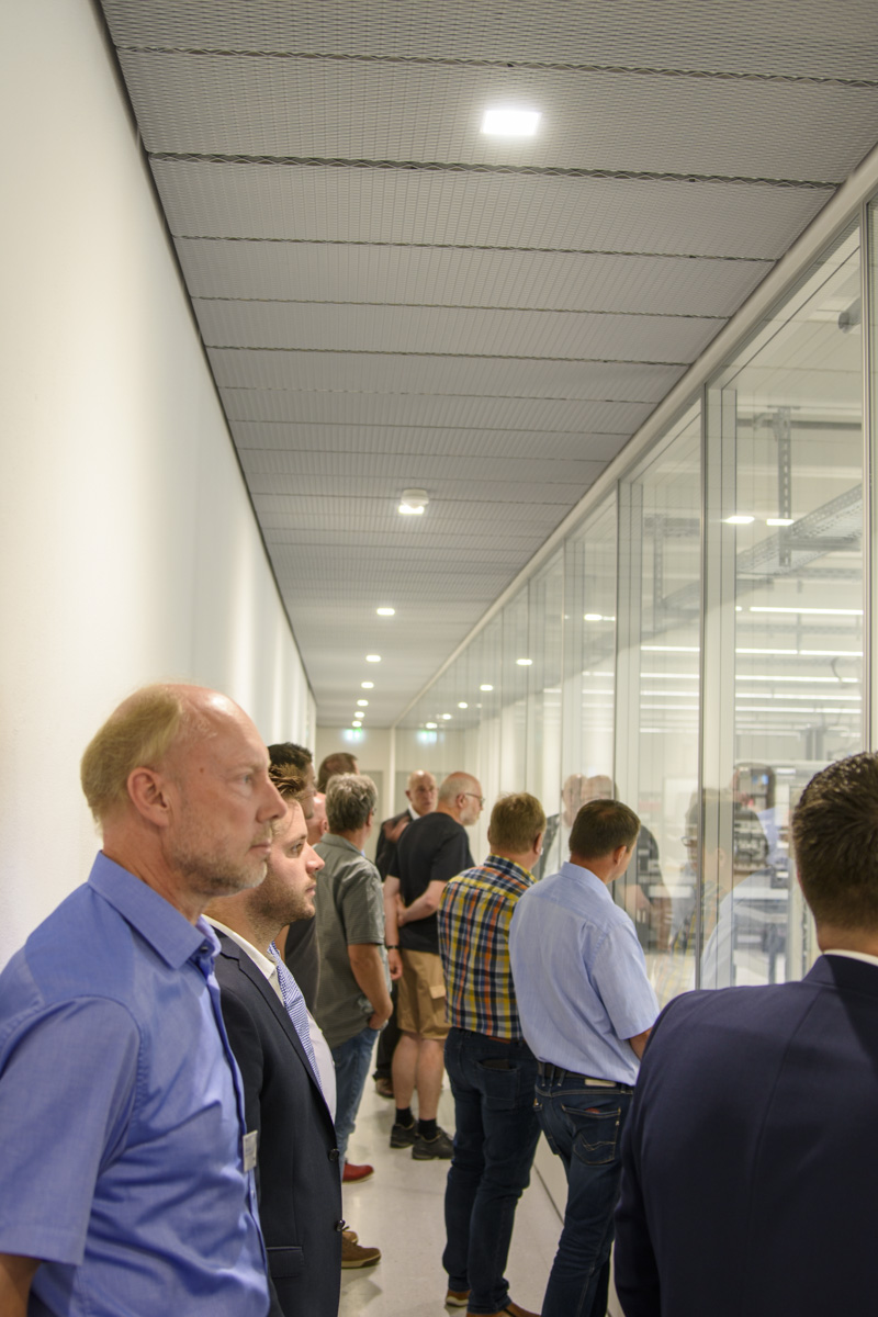 A group of people stands in a corridor, facing large glass windows that showcase an industrial setting, engaged in conversation and observation.