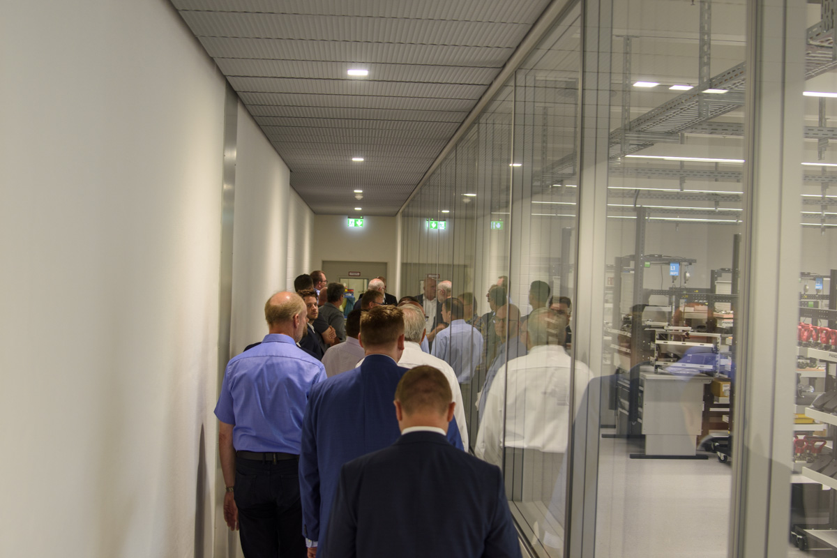 A group of people walking down a hallway, viewed from behind, with glass walls revealing machinery or equipment in the background.