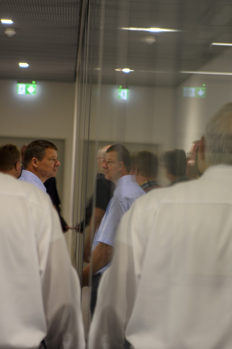 A group of people in formal attire stands behind a glass partition, with reflections creating a layered effect. Some individuals engage in conversation while others observe.