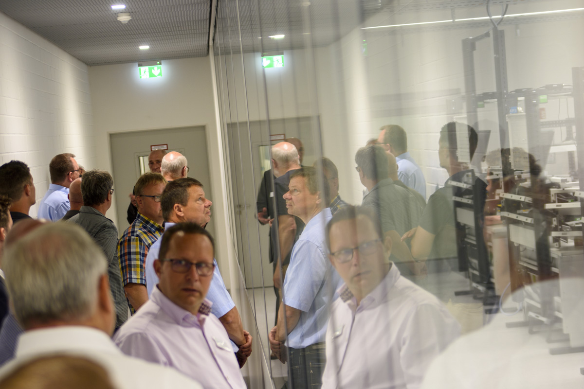 A group of people stands in a hallway, looking towards a glass enclosure filled with equipment. Reflections and expressions suggest interest in the showcased items.